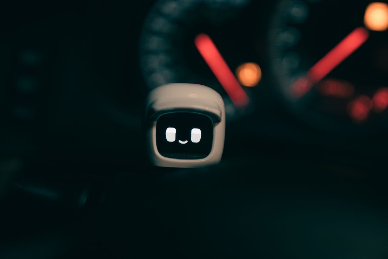 A small robotic figure with a smiling face on a car dashboard, lit by blurred speedometer lights at night.